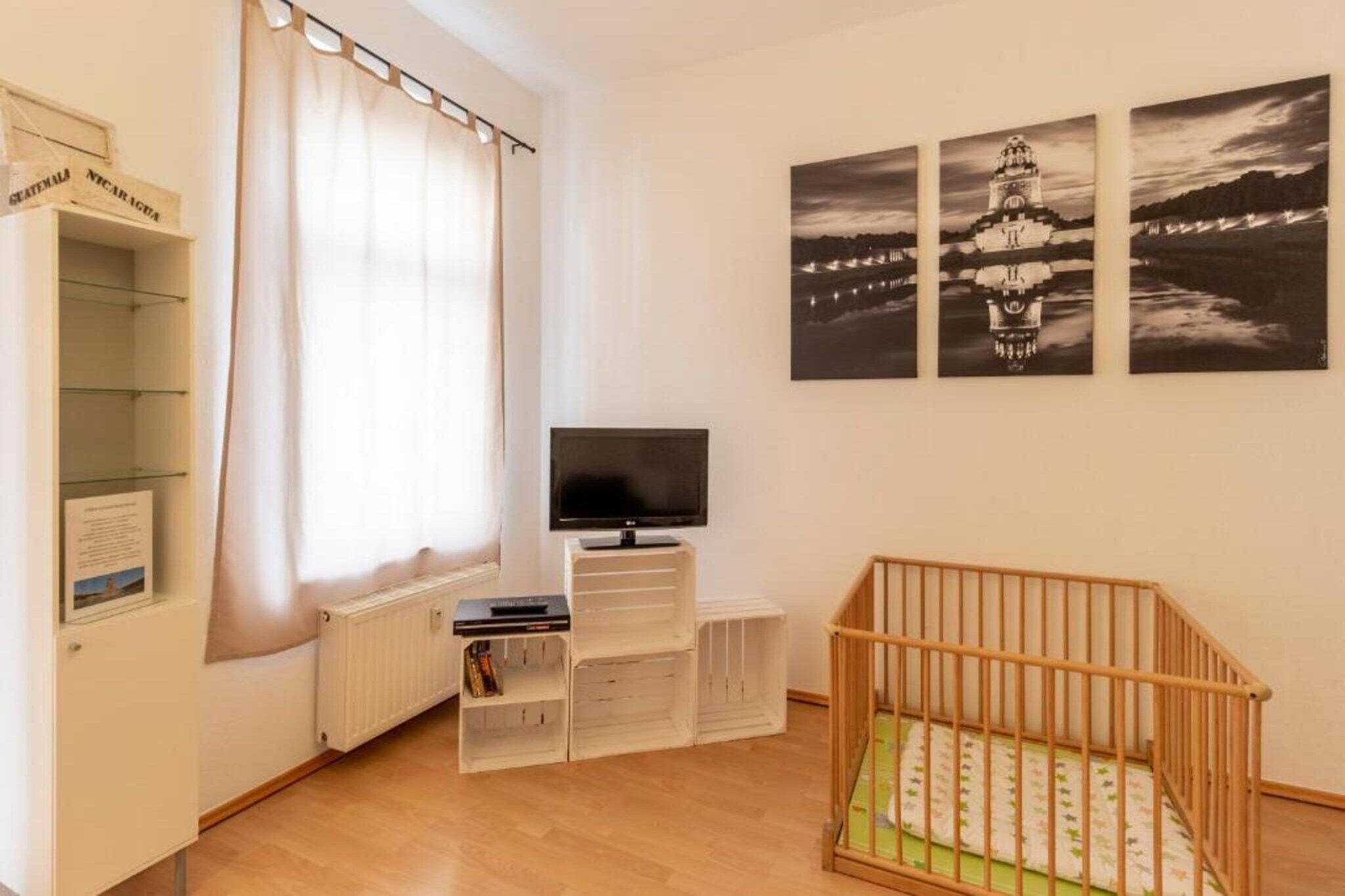 Ein Zimmer mit einem Kinderbett, einem Fernseher auf einem Beistelltisch, einem Fenster mit Vorhängen und drei Schwarz-Weiß-Fotos an der Wand.