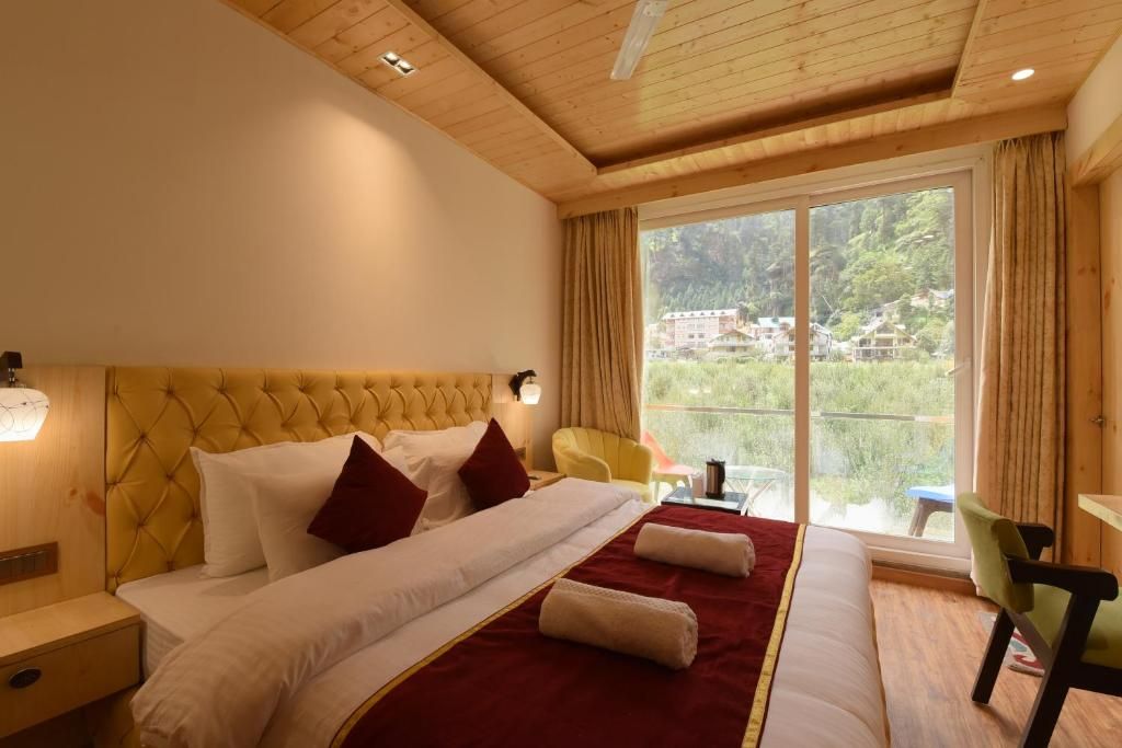 A hotel room with a large bed, a view of the outdoors, a wooden ceiling, and a small table with a chair.