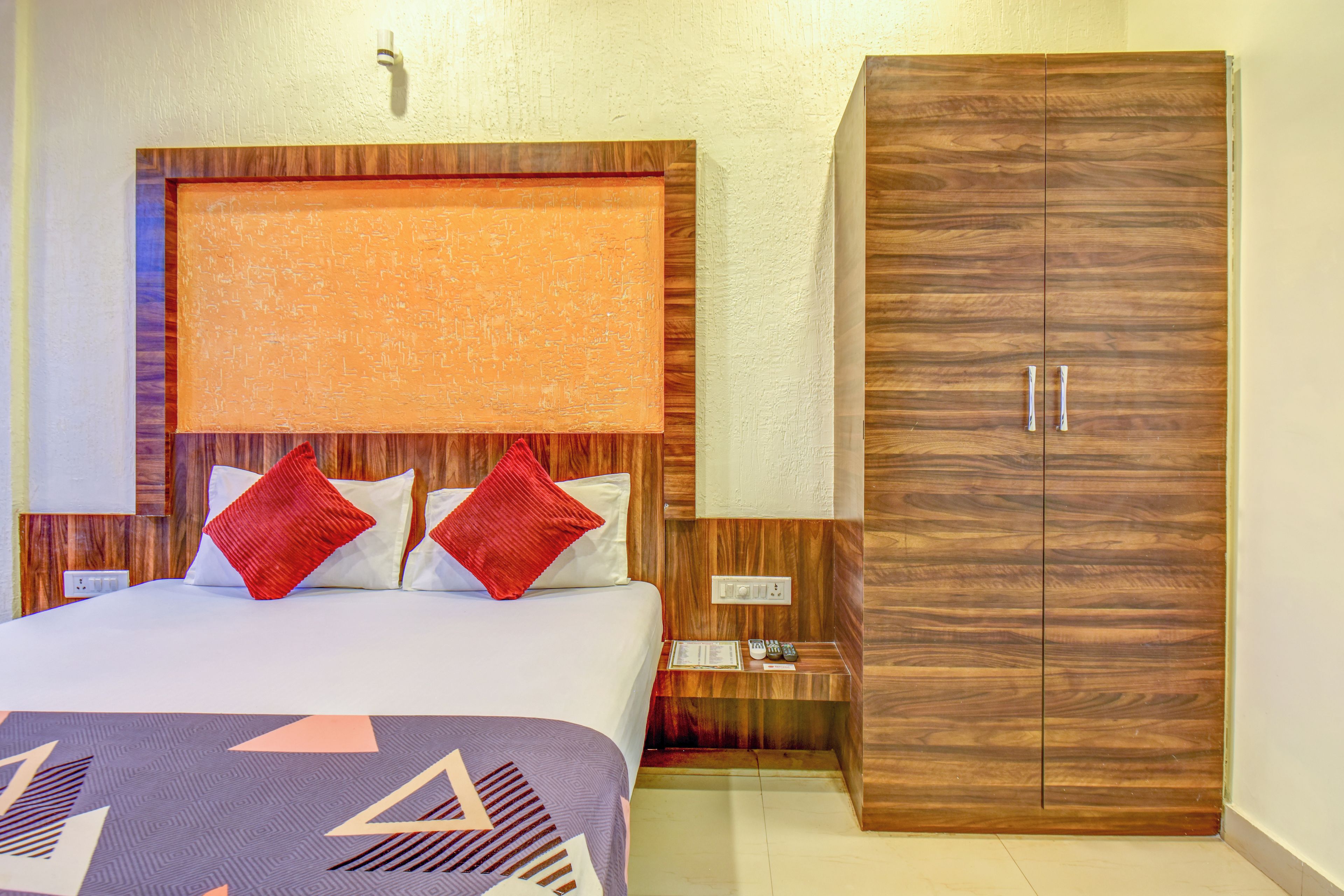 A hotel room with a bed, two red pillows, a nightstand, a wooden wardrobe, and a wall-mounted light fixture.