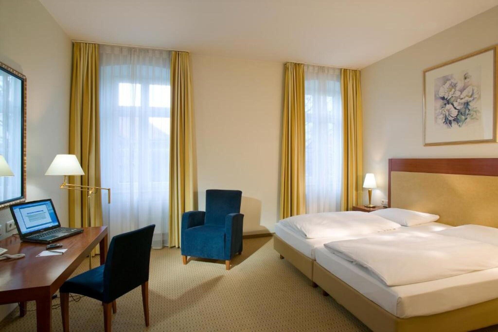 A hotel room with a desk, two chairs, a bed, and a painting on the wall.