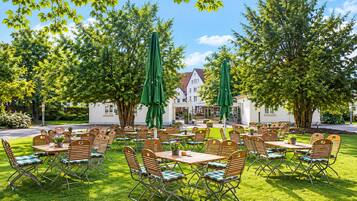 An outdoor seating area with tables and chairs, surrounded by trees and a building in the background.