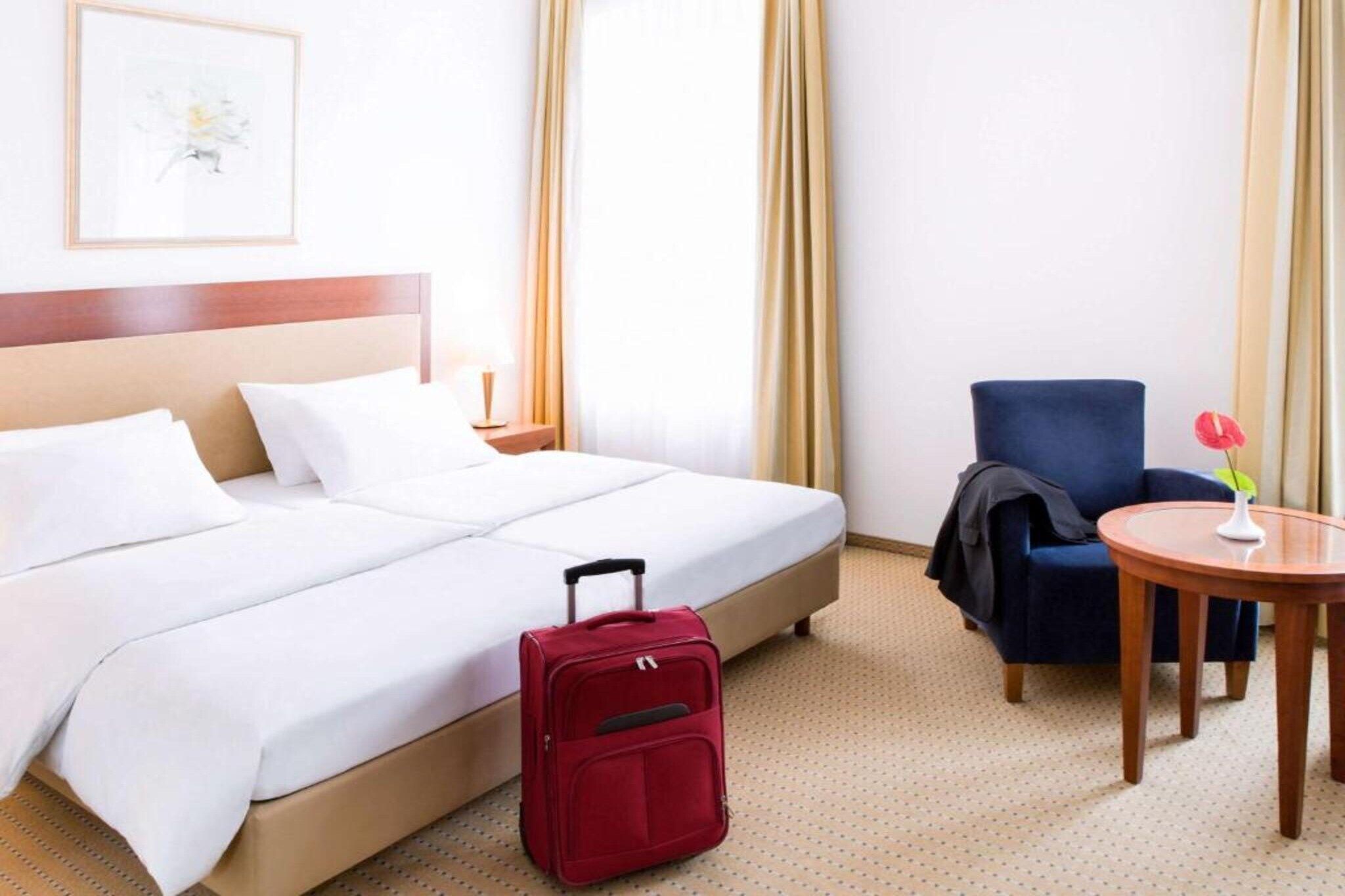 Ein Hotelzimmer mit einem Bett, einem roten Koffer, einem blauen Sessel, einem kleinen runden Tisch und einem an der Wand hängenden Bild.