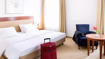 Ein Hotelzimmer mit einem Bett, einem roten Koffer, einem blauen Sessel, einem kleinen runden Tisch und einem an der Wand hängenden Bild.
