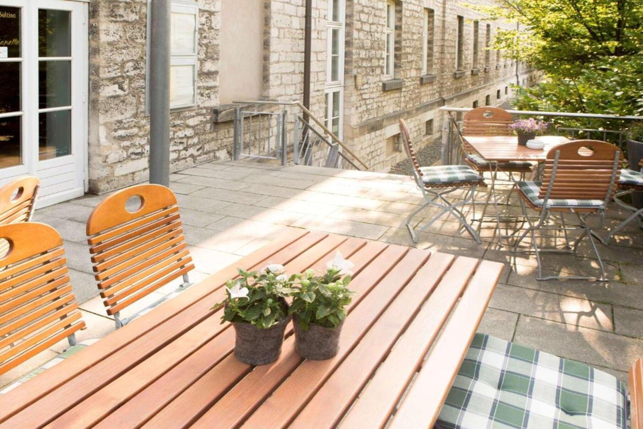Eine Terrasse im Freien mit Holztischen und -stühlen, eine kleine Gartenanlage mit Blumentöpfen und ein Steinhaus im Hintergrund.