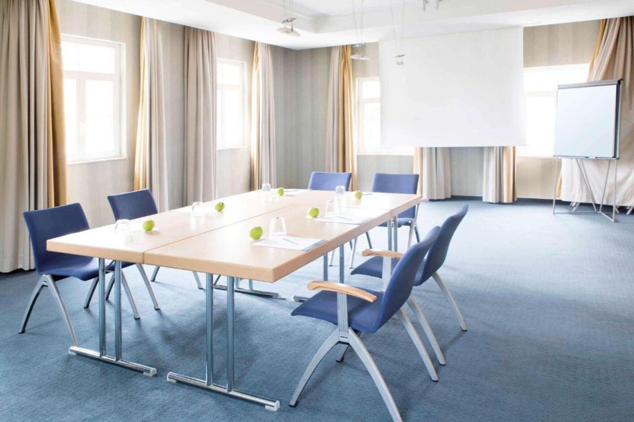 A conference room with a long table, blue chairs, a whiteboard, and a projector screen.