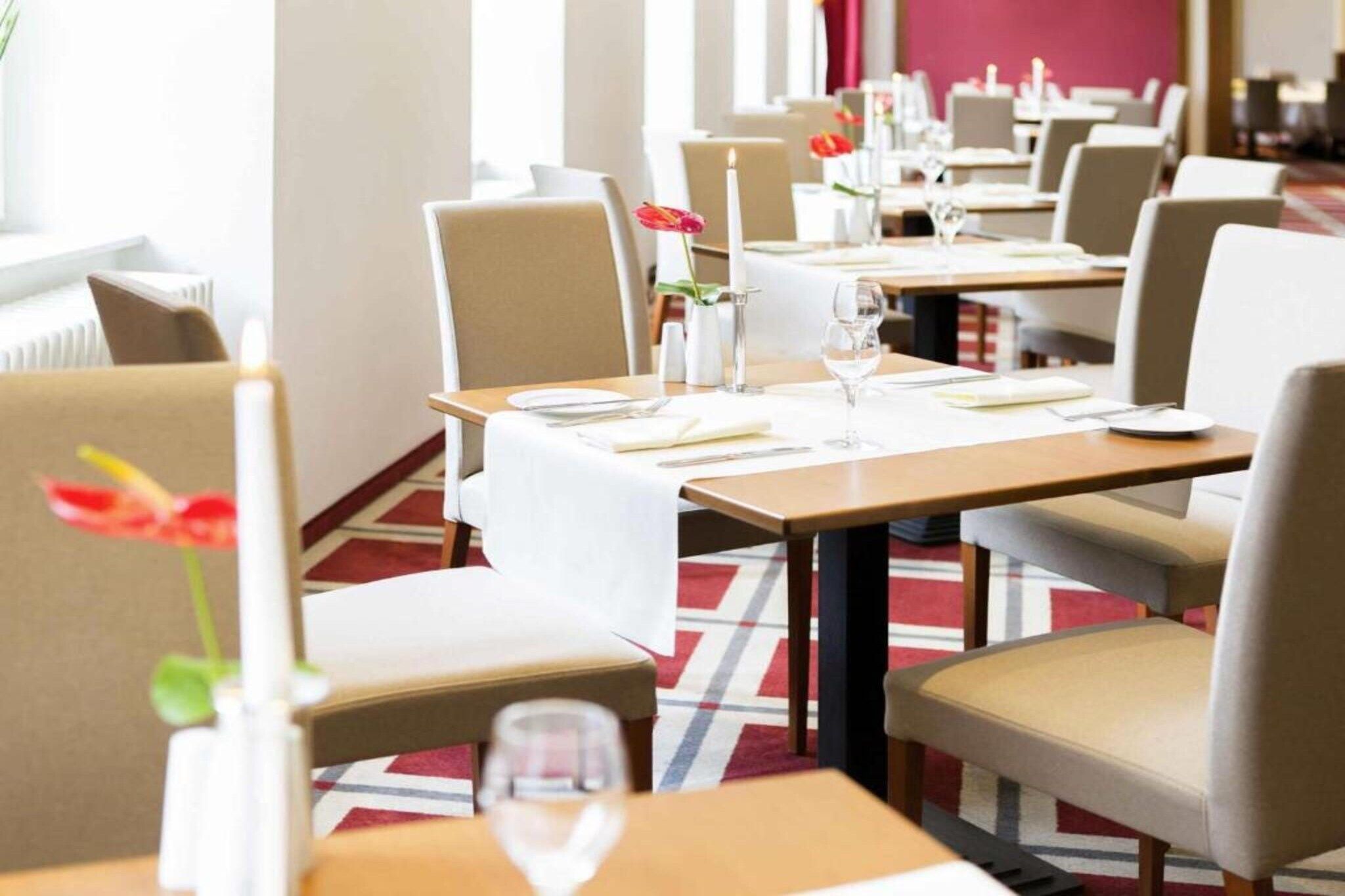 Une salle à manger d’hôtel avec des tables préparées pour deux personnes, des chaises et un vase contenant une seule fleur.
