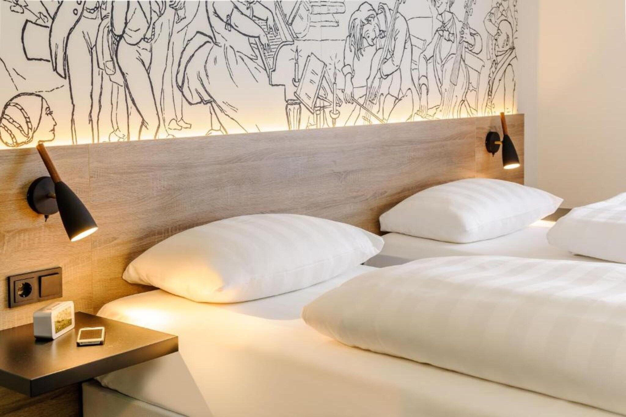 Ein Hotelzimmer mit einem hölzernen Kopfteil, zwei Nachttischlampen, einem Nachttisch mit einer Uhr und einer Fernbedienung sowie zwei weißen Kissen.