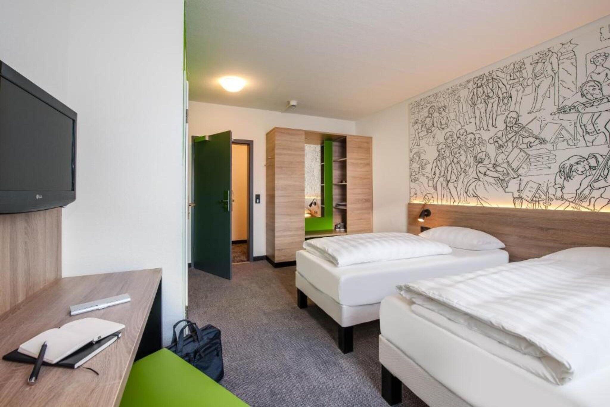 Ein Hotelzimmer mit zwei Betten, einem Schreibtisch, einem Sessel, einem Fernseher und einer Wand mit einem Wandgemälde.