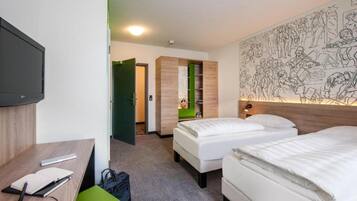 Ein Hotelzimmer mit zwei Betten, einem Schreibtisch, einem Sessel, einem Fernseher und einer Wand mit einem Wandgemälde.