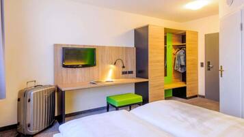 A hotel room with a bed, a desk with a flat-screen TV, a suitcase, a green stool, and a walk-in closet.