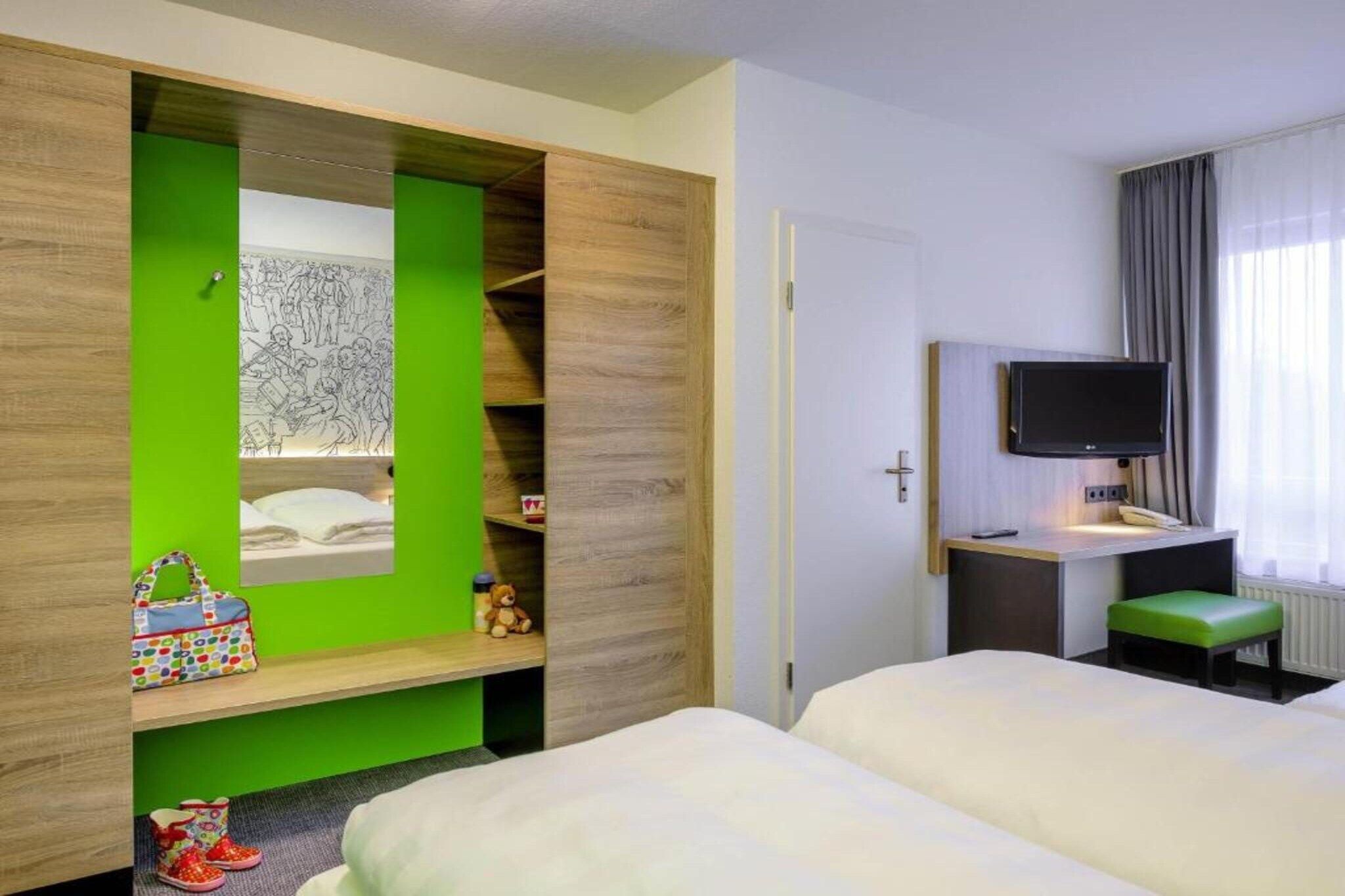 A hotel room with two beds, a desk with a flat-screen TV, a green accent wall, and a window with curtains.