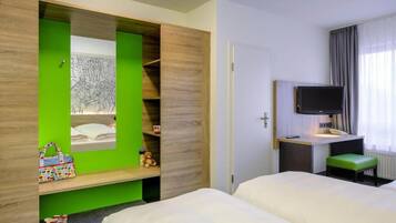 A hotel room with two beds, a desk with a flat-screen TV, a green accent wall, and a window with curtains.