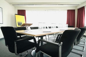 A conference room with a long table, black chairs, a projector screen, and a window with curtains. - Sunday Hotel Halle Zentrum - Suite (Halle (Saale))