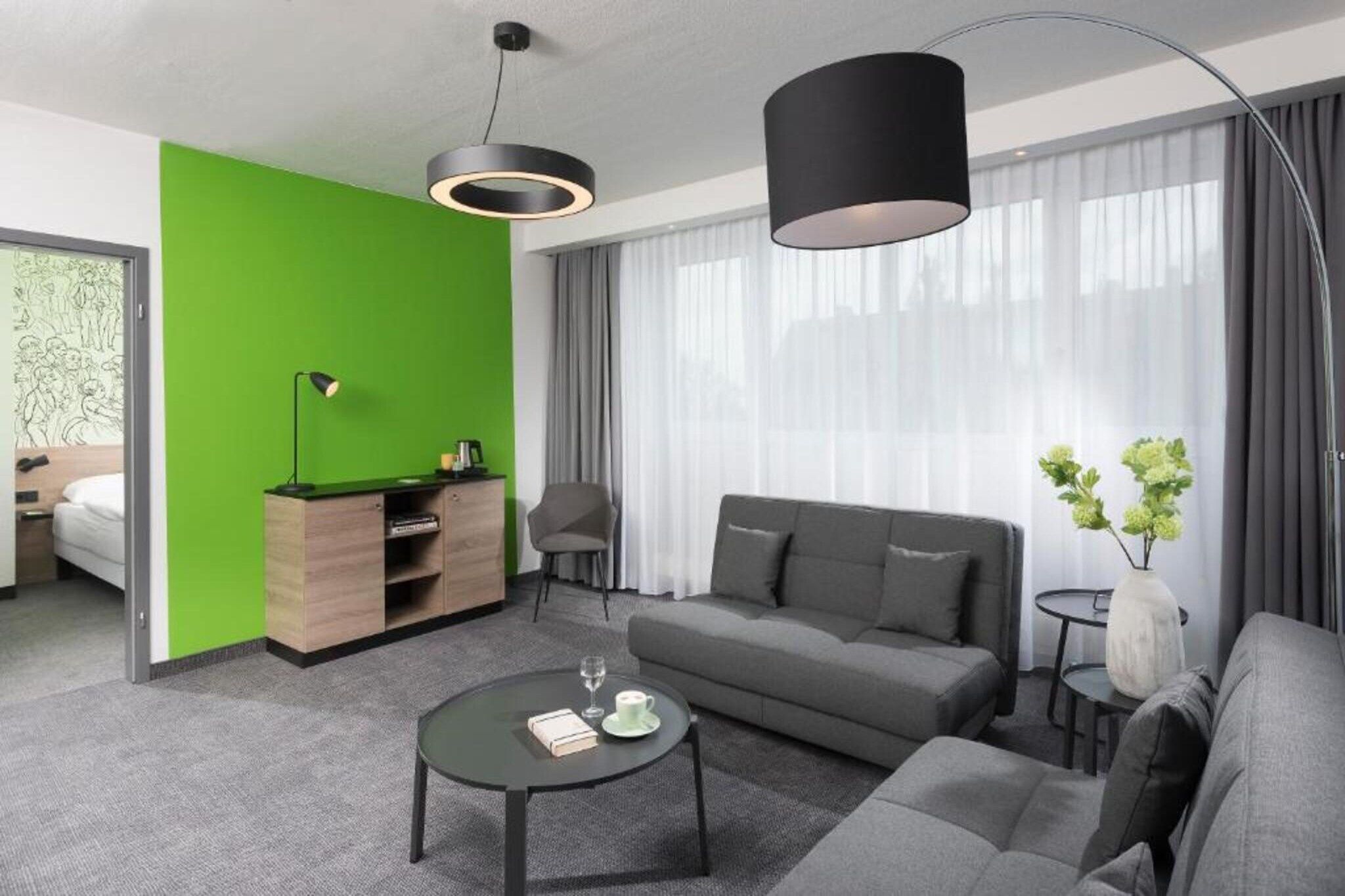 Ein modernes Wohnzimmer mit grüner Akzentwand, einem grauen Sofa, einem Couchtisch und einer schwarzen Pendelleuchte.