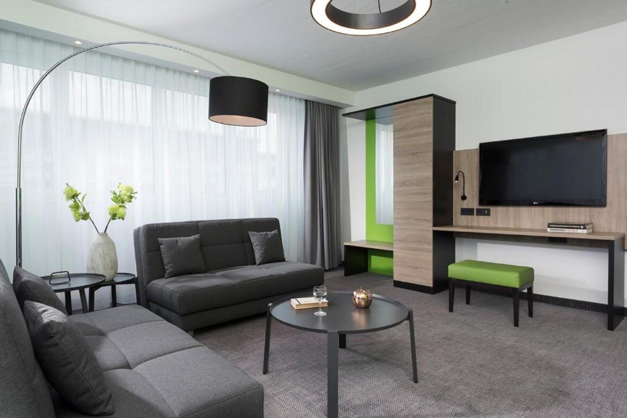 Ein modernes Hotelzimmer mit einer Couch, einem runden Couchtisch, einem Fernseher und einer grünen Bank.