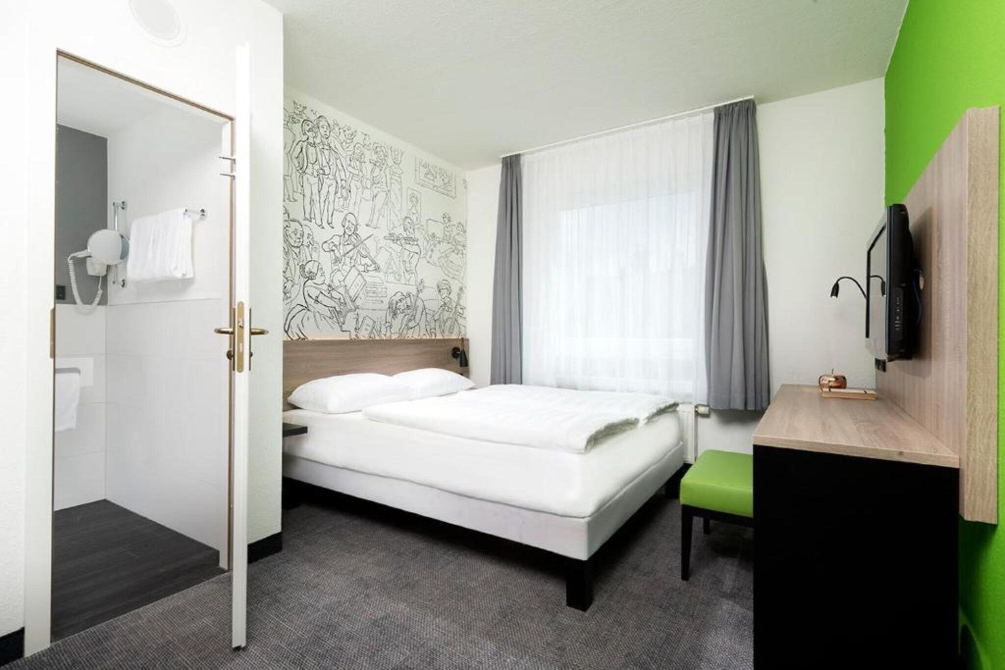 A hotel room with a bed, a desk, a mirror, a wall with a mural, and a bathroom visible through an open door.