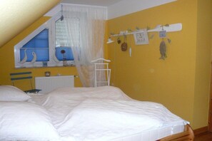 A bedroom with a sloped ceiling, a bed with white bedding, a window with blue shutters, and a wall-mounted shelf with decorative items. - Ulferts am Timmeler Meer (Großefehn \/ Timmel)