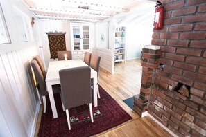 A dining area with a white table, brown chairs, a red patterned rug, a brick fireplace, and a white cabinet with glass doors. - Fresh Water Fjord Cabin (Agder)