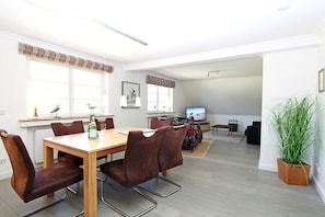 A dining area with a wooden table and brown chairs, a television in the background, and a potted plant. - Holiday apartment with 3 bedrooms (Hörnum \/ Sylt)