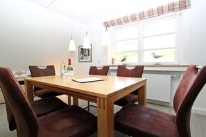 A dining area with a wooden table and six chairs, a window with a view of birds, and a radiator below the window. - Holiday apartment with 3 bedrooms (Hörnum \/ Sylt)