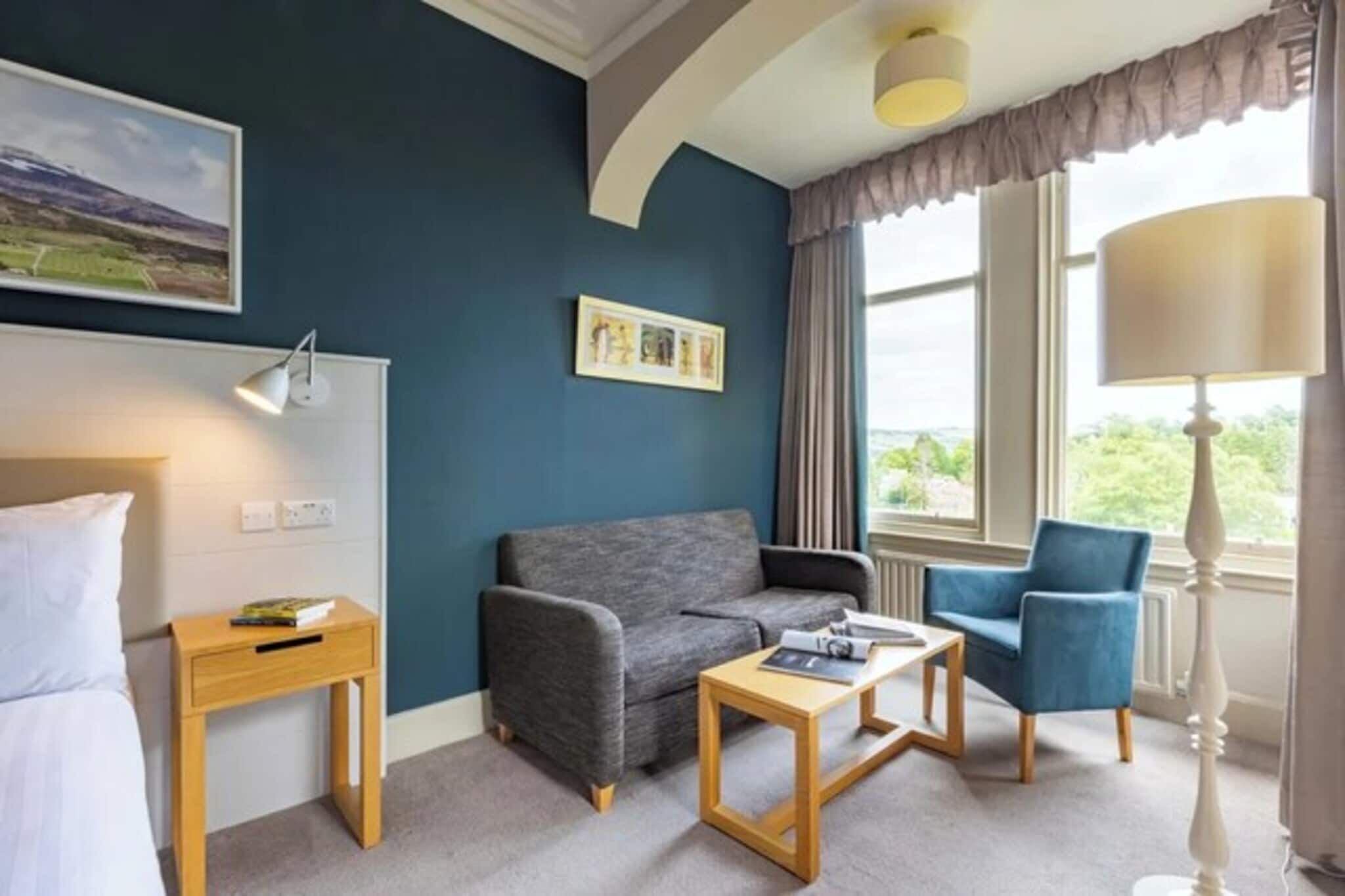 A hotel room with a bed, a grey sofa, a blue armchair, a wooden side table, and a window with curtains.