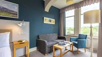A hotel room with a bed, a grey sofa, a blue armchair, a wooden side table, and a window with curtains.
