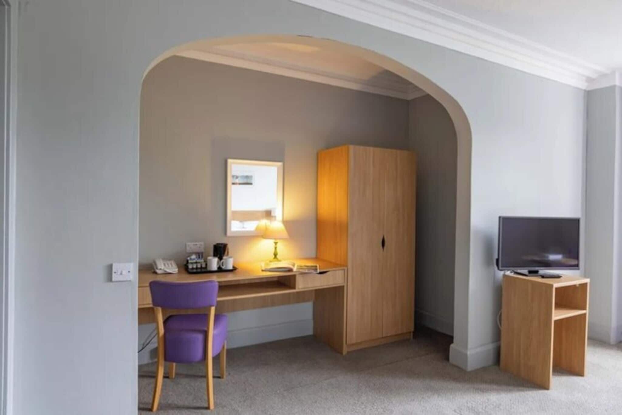 A compact room with a desk, chair, wardrobe, television, and a small shelf.