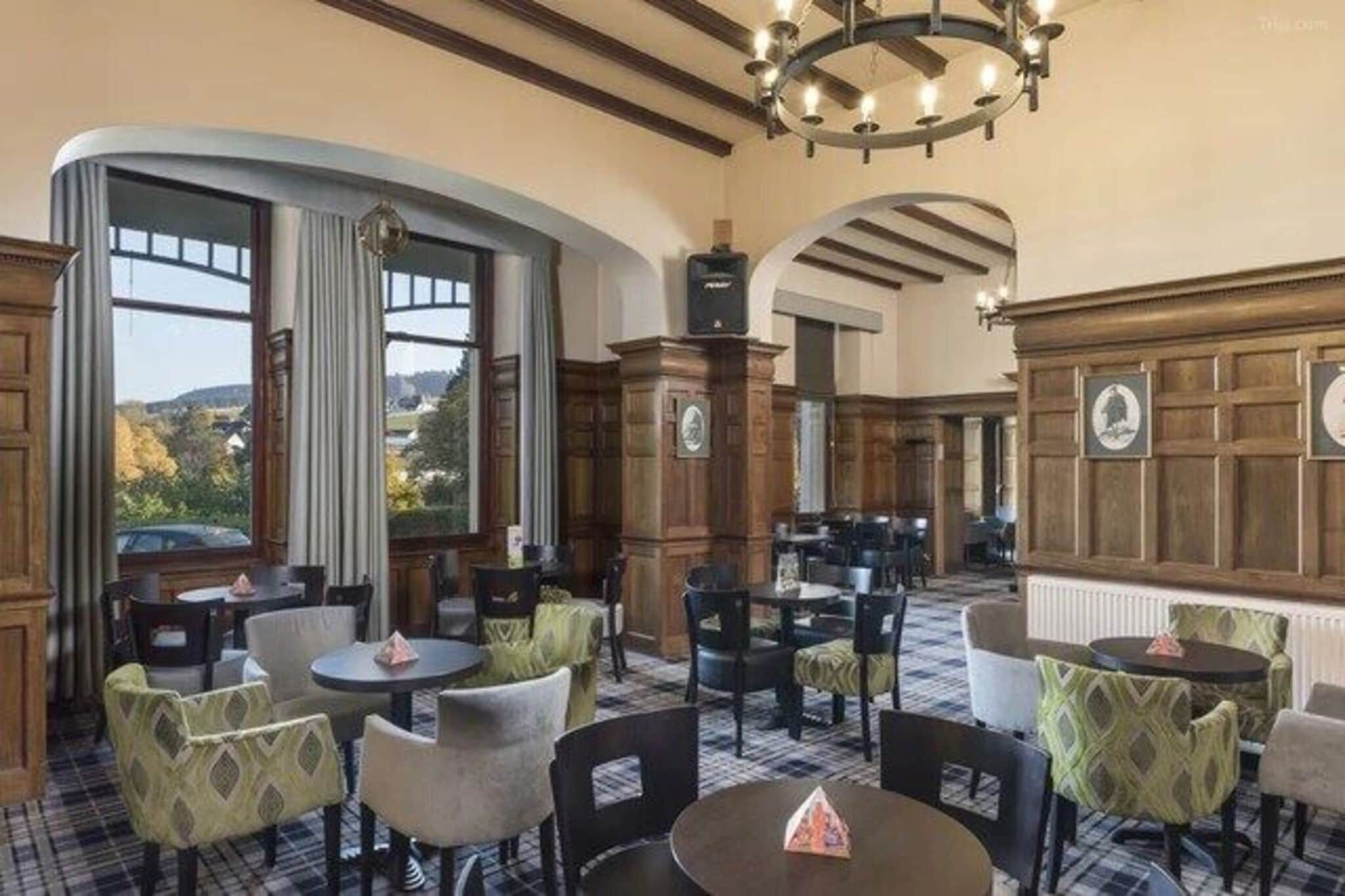 A hotel dining area with round tables and chairs, wooden paneling, and large windows offering a view.