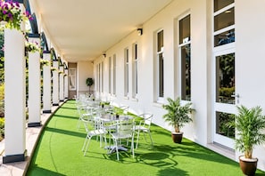 Een overdekt buitengebied met rijen tafels en stoelen, potplanten en een vloer van kunstgras.
