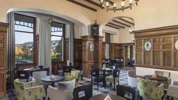 A hotel dining area with round tables and chairs, wooden paneling, and large windows offering a view.