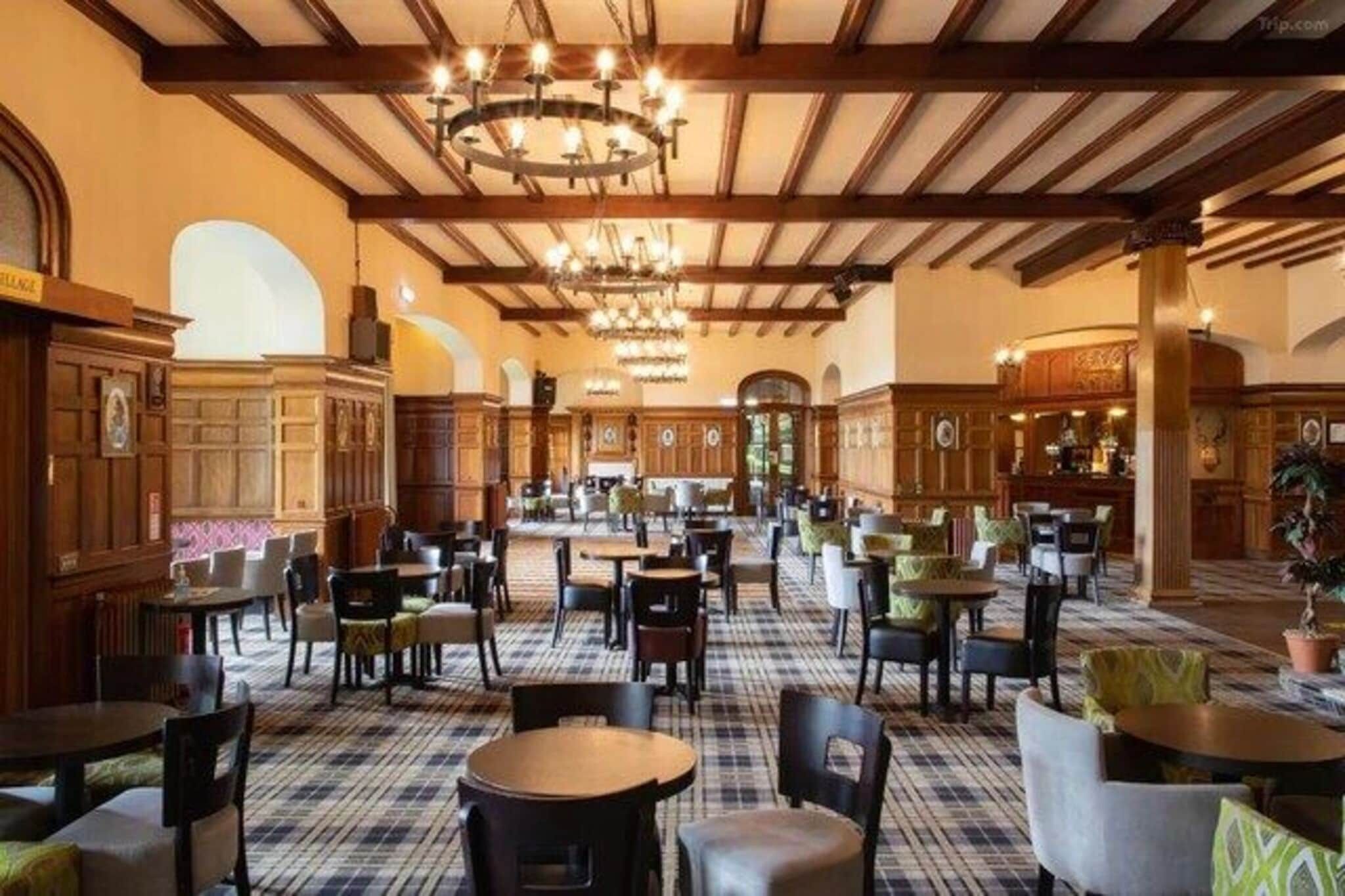 A spacious dining area with multiple tables and chairs, wooden paneling, and a high ceiling with chandeliers.