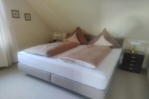 Een net opgemaakt bed met kussens en een deken, een nachtkastje met een lamp, en een klein dressoir.