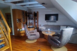 A loft-style living area with a wooden staircase, a grey sofa, a recliner, a small round table, and a bookshelf with decorative items. - Holiday apartment with 1 bedroom (Jemgum-Ditzum)