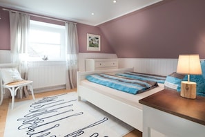 A bedroom with a bed, a chair, a bedside table, a lamp, and a window with curtains. - Holiday home and Lootsje (Borkum)