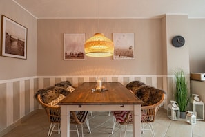 A dining area with a wooden table, wicker chairs, a hanging light fixture, framed pictures on the wall, a clock, and a potted plant. - Holiday apartment in Binz with swimming pool (Binz)