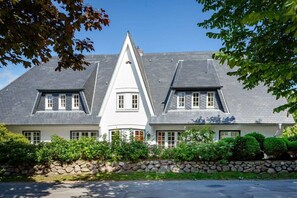 Ein zweigeschossiges Haus mit einem steilen Satteldach, einem zentralen weißen Turm und einer Steinmauer davor.