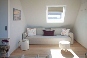 Une pièce confortable avec un canapé, un petit pouf rond, une fenêtre avec des stores et un tableau au mur.
