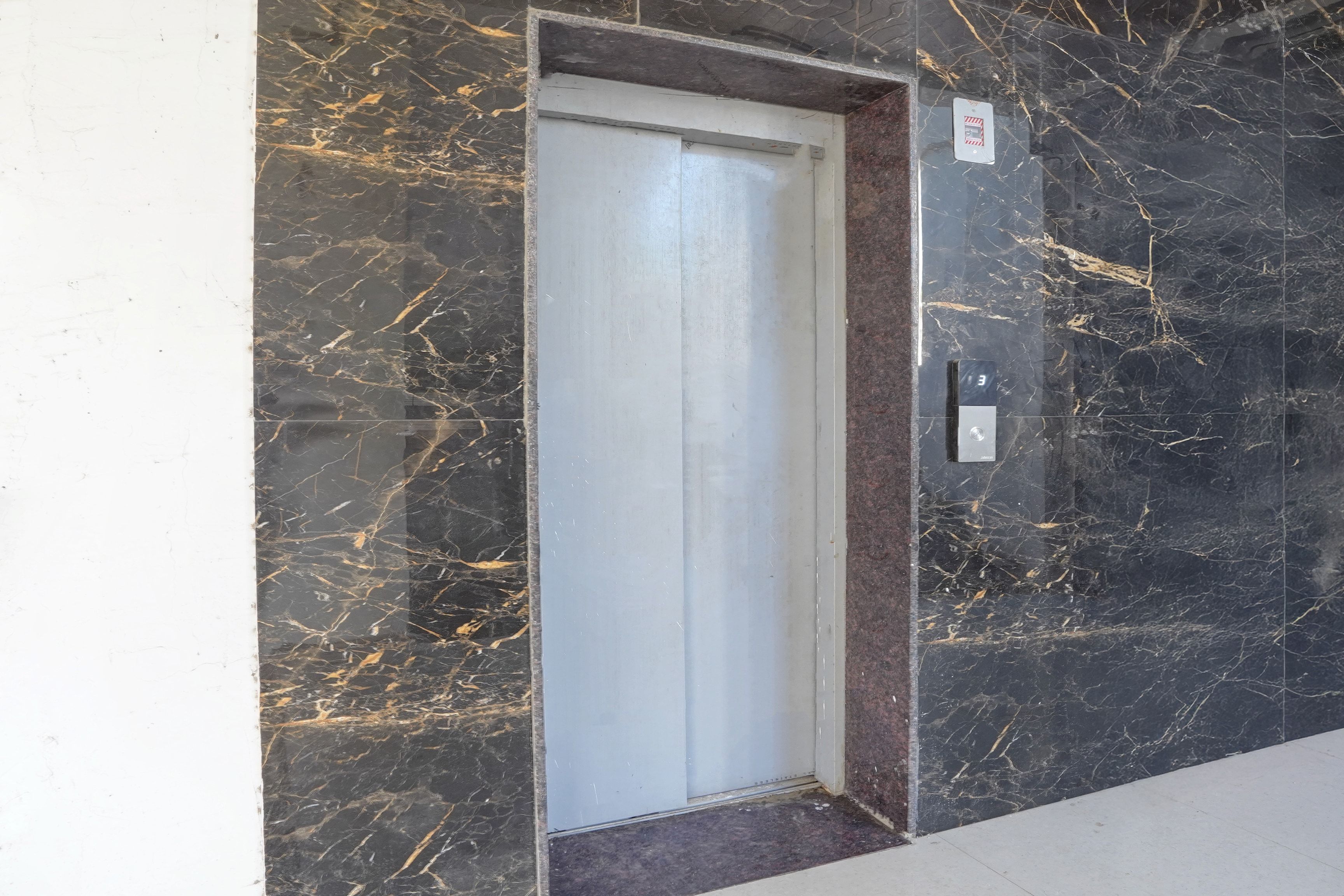 A closed elevator door with a marble wall and a fire alarm panel to the right.