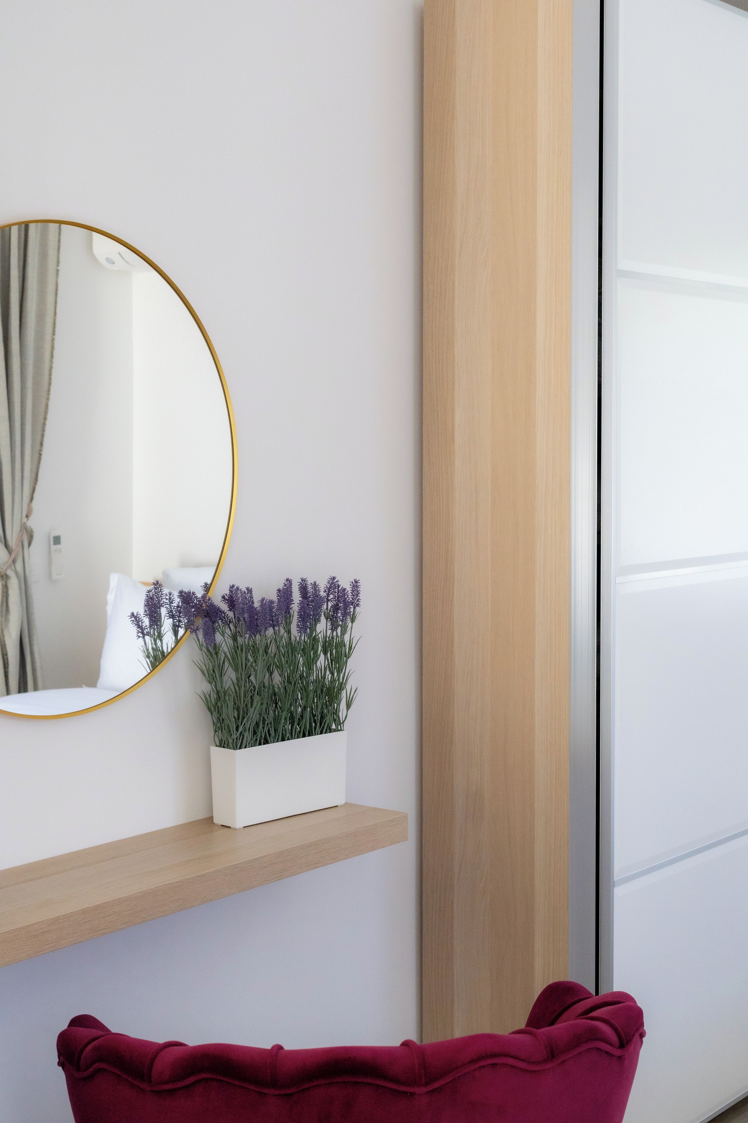 Une pièce avec un mur blanc, un miroir rond doté d’un cadre doré, une étagère blanche sur laquelle est posée une plante de lavande en pot, et une porte en bois.
