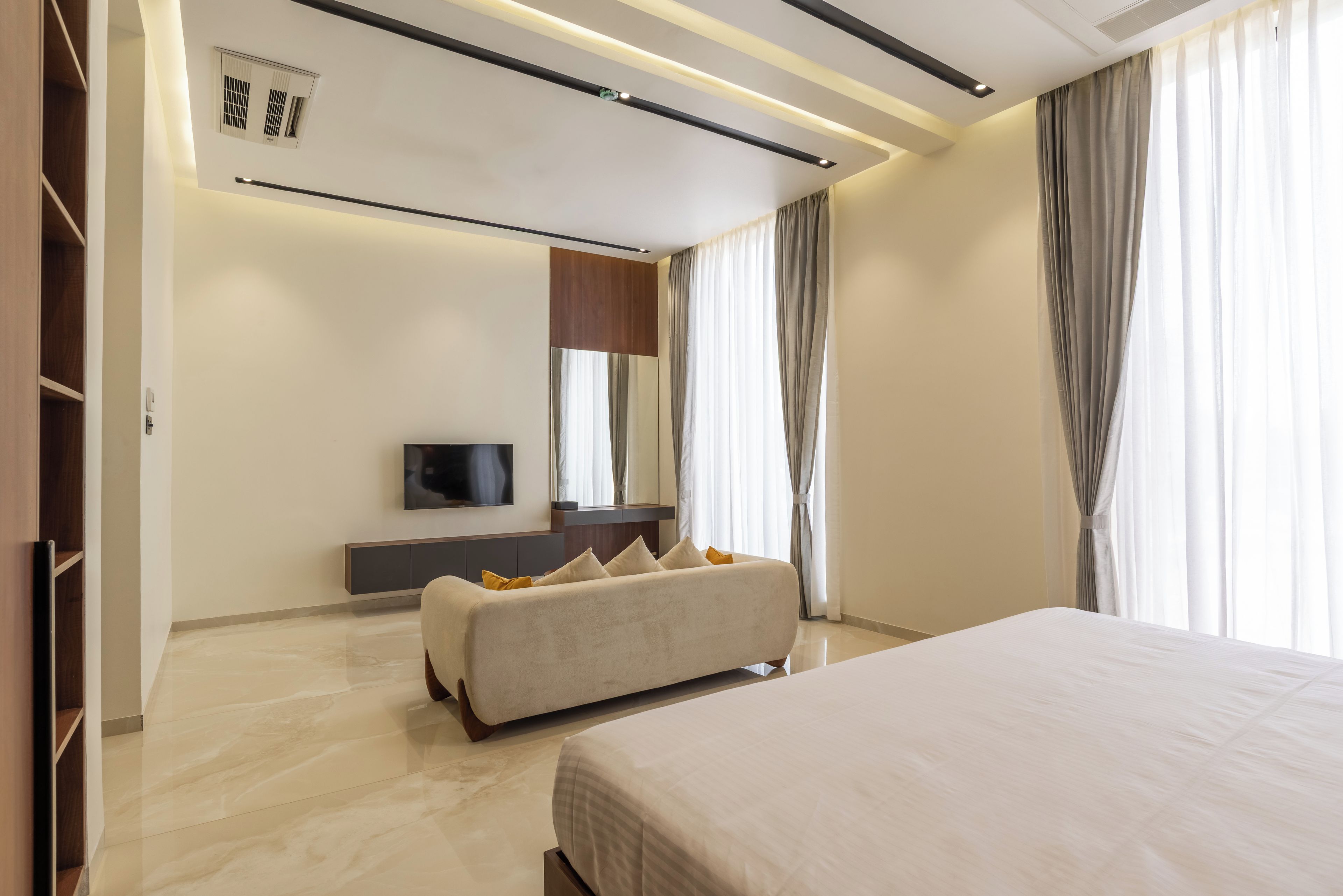 Un dormitorio moderno con cama, sofá, televisor y un amplio ventanal con cortinas.