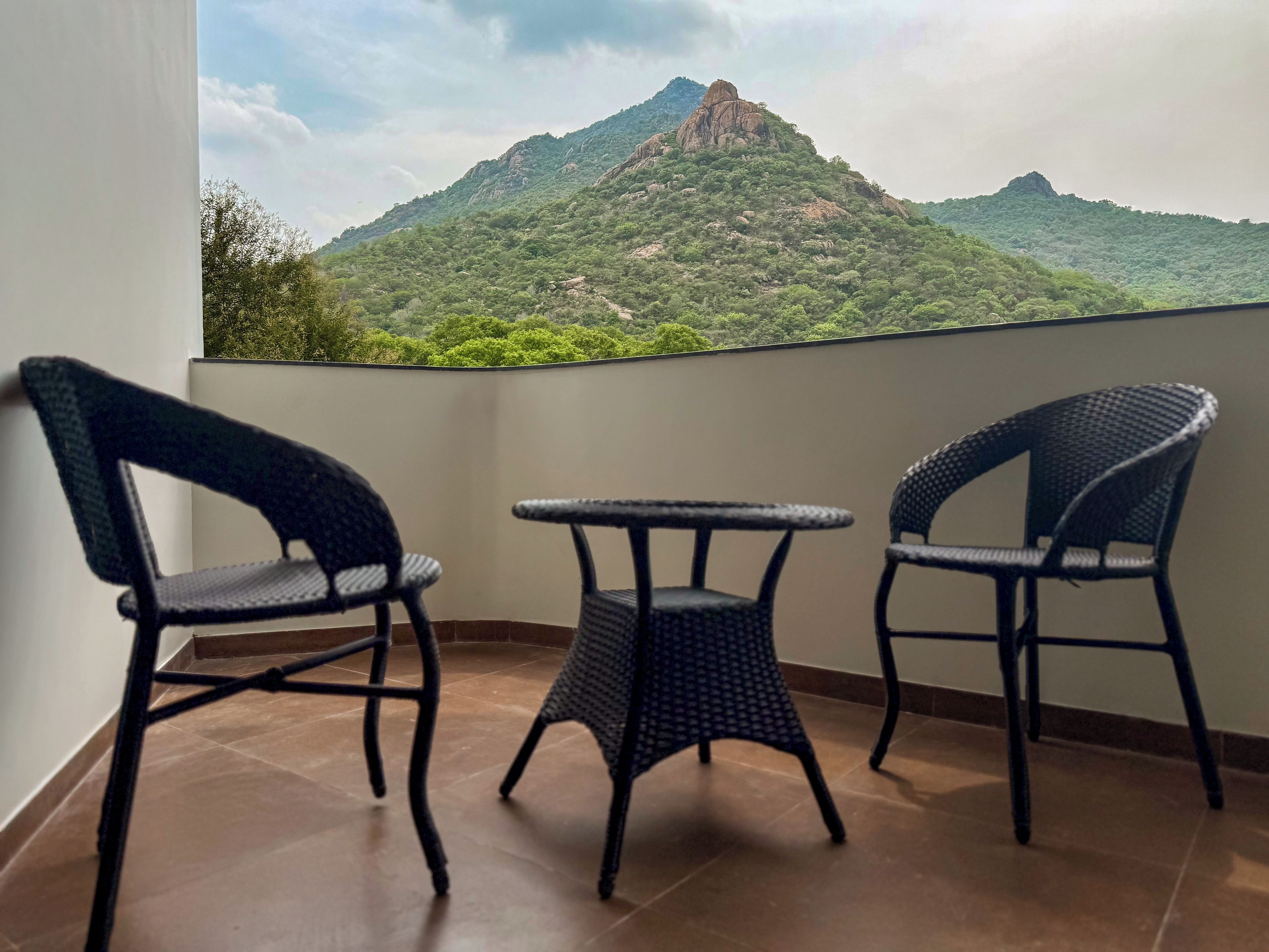 Un balcón con tres sillas de mimbre y una pequeña mesa, con vistas a un paisaje montañoso.