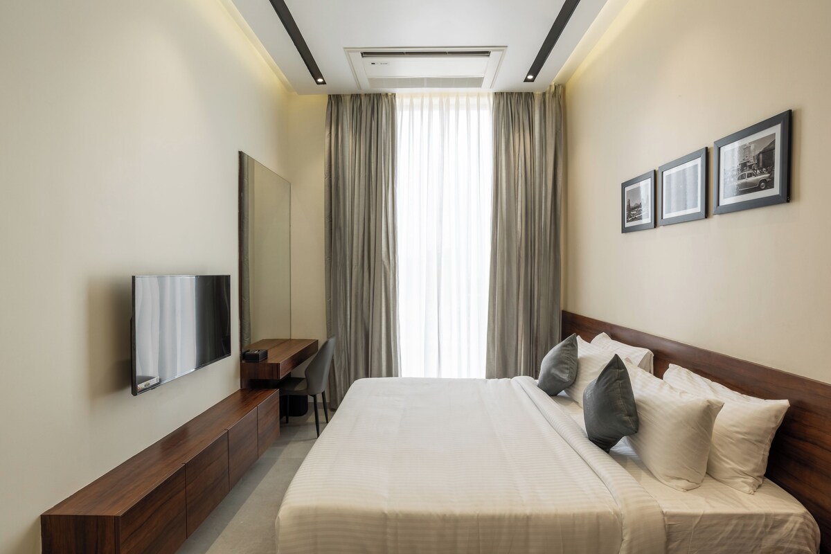 Una camera d'albergo con un letto, una scrivania, una sedia, una TV e una finestra con tende.