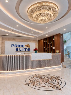 Un hall d'accueil d'hôtel au design moderne, agrémenté d'un grand lustre, d'un comptoir d'accueil sur lequel est apposé un panneau indiquant « PRIDE ELITE », et d'un motif décoratif sur le sol.