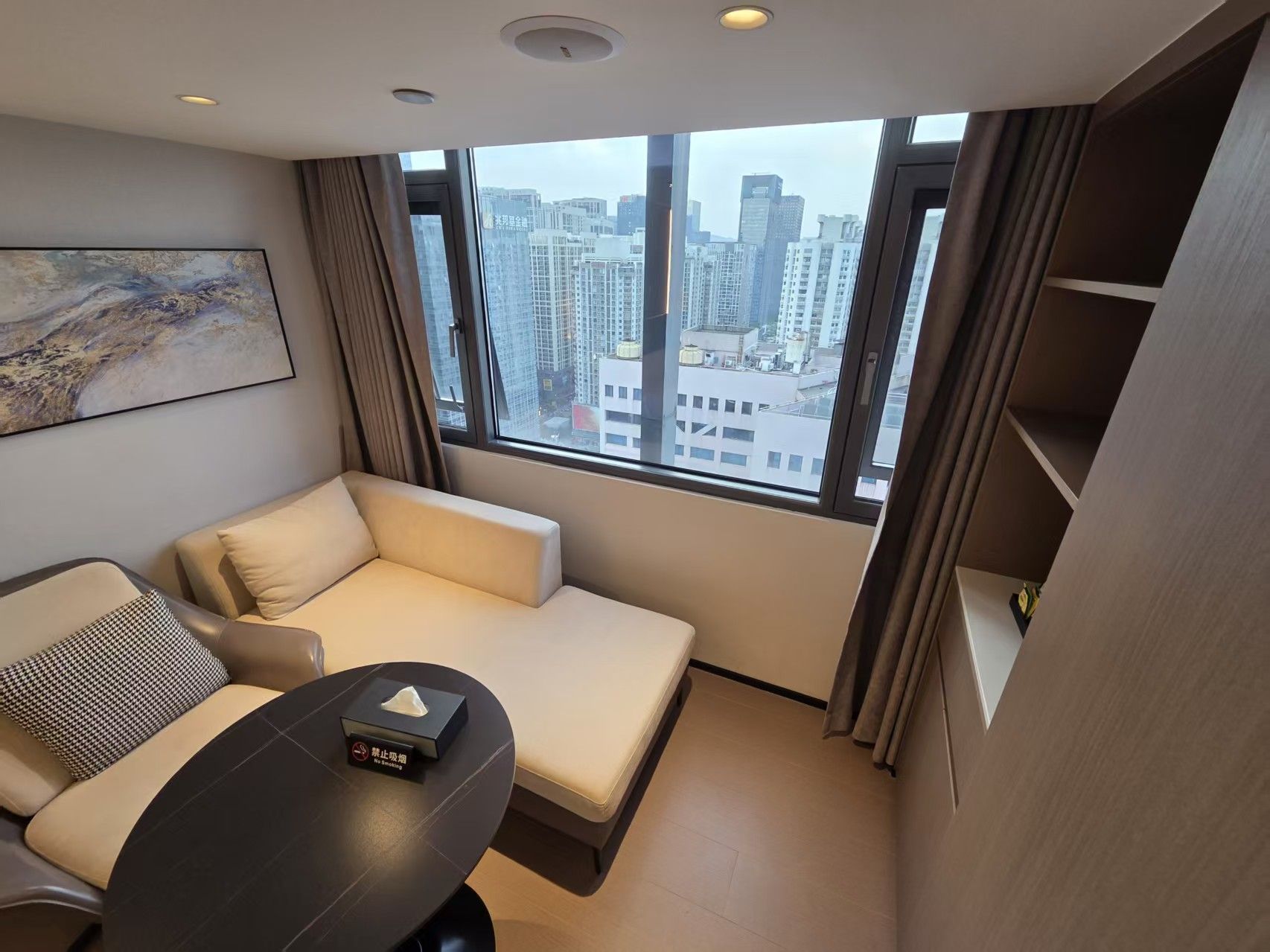 A modern hotel room with a sofa, a small table, and a view of the cityscape through large windows.