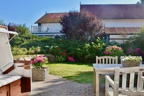 Une terrasse avec une table et des chaises en bois, un panier en osier et un jardin où poussent différentes plantes et fleurs.