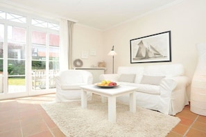 A white furnished room with a large window, a framed picture on the wall, and a small table with fruit. - at the Arnfried country house (Wenningstedt)