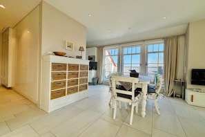 A spacious living area with a dining table, chairs, a white cabinet with drawers, a television, and large windows. - Holiday apartment with 2 bedrooms (Norderney)