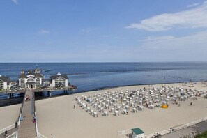 Ein Strand mit Reihen weißer Strandsonnenschirme und einem Steg, im Hintergrund sind Gebäude zu sehen.