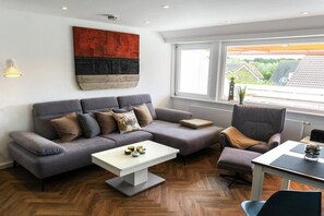 A modern living room with a grey sofa, a white coffee table, and a view of houses through large windows. - Holiday apartment with 1 bedroom (Norderney)