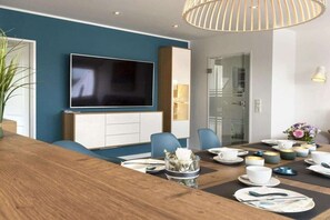 A modern dining area with a wooden table set for a meal, a flat-screen TV mounted on the wall, and a glass door leading to another room. - Holiday apartment with 1 bedroom (Norderney)